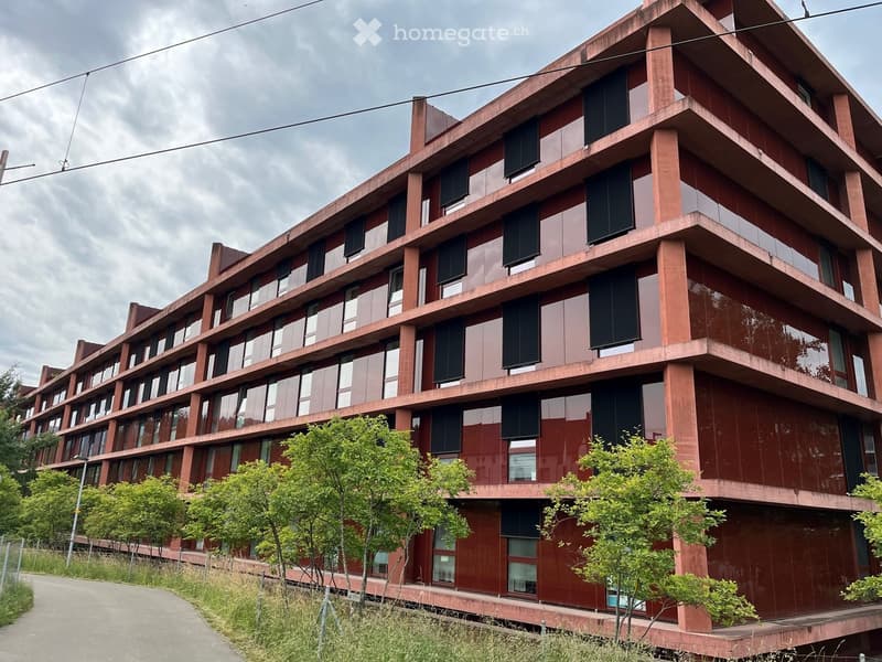 Modern multi-story building with red brick facade, large windows, and balconies. Surrounded by greenery and a paved pathway.