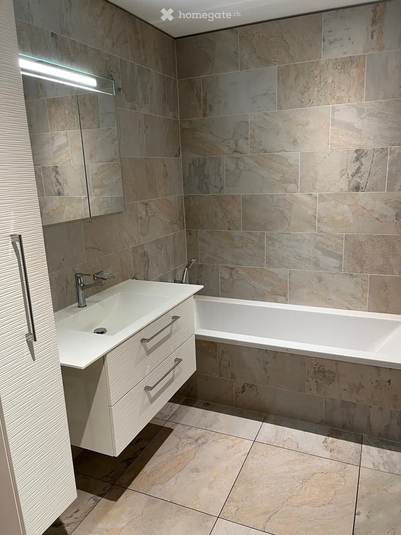 Modern bathroom with marble tiles, a white sink with a modern faucet, a bathtub, and a large mirror with a built-in light fixture.