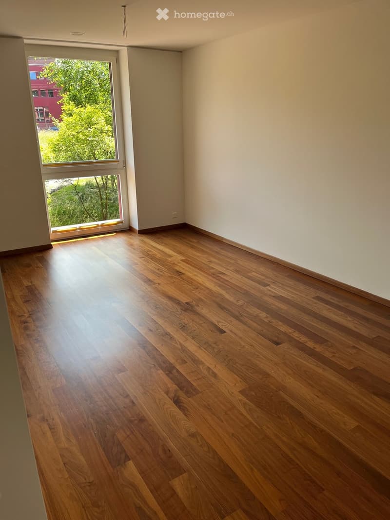 Empty room with wooden flooring and large window letting in natural light. Bare walls and no furniture.