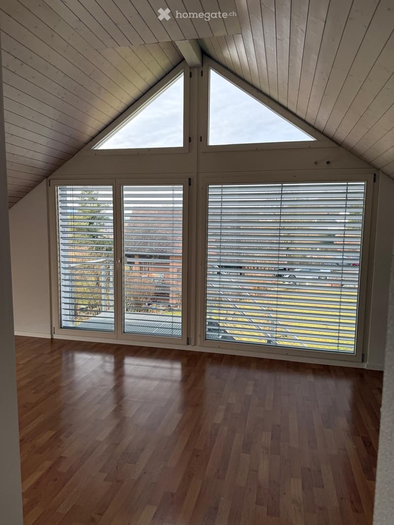 Bright, airy attic room with wooden floors, large windows, and skylights, featuring a sloped wooden ceiling and modern blinds.