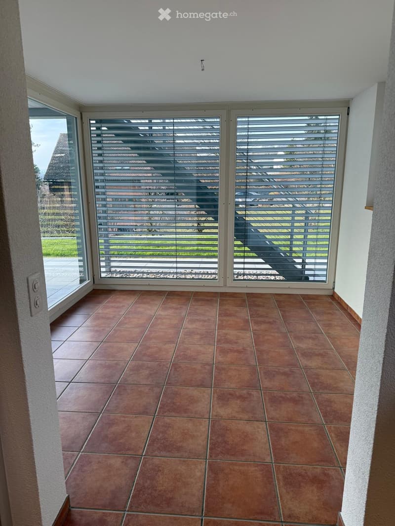 Empty room with brown tiled floor and large windows with blinds, leading to an outdoor area.
