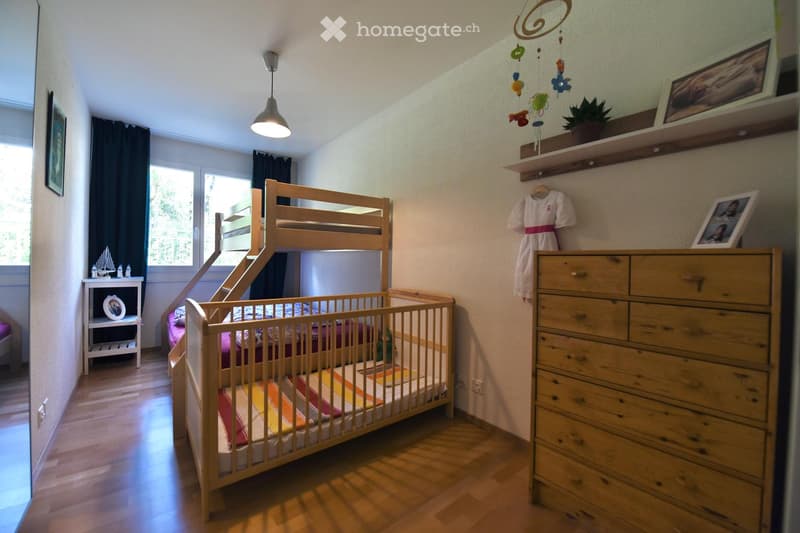 A well-lit bedroom with a bunk bed, a crib, a wooden dresser, shelves, and a small table with a picture frame. The room features wooden flooring and large windows with dark curtains.