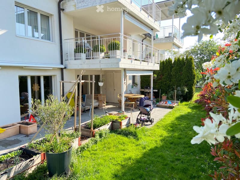 Well-maintained garden with green lawn, potted plants, raised garden beds, outdoor furniture including a table and chairs, and a stroller near the patio area.