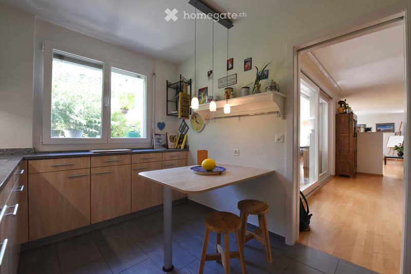 Bright kitchen with wooden cabinets, a central island table, two wooden stools, and a window overlooking greenery. Shelves hold decorative items and a clock.