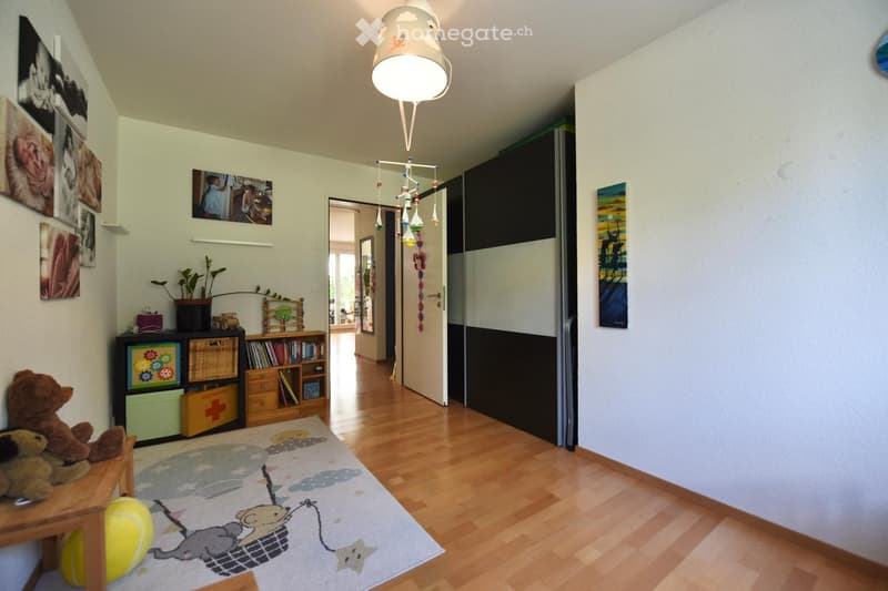 A well-lit hallway with wooden flooring, a colorful rug, a small table with a teddy bear, a bookshelf, and a hanging mobile with toys.