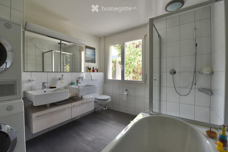 Modern bathroom with double sinks, a bathtub, a toilet, a large mirror, a shower area with a glass enclosure, and a window offering a view of greenery. The space includes a cabinet under the sinks and a towel rack near the shower.