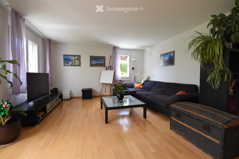 Modern living room with light wooden flooring, large windows with curtains, a dark blue sectional sofa, a glass coffee table, a TV on a black stand, and several decorative plants and artwork.