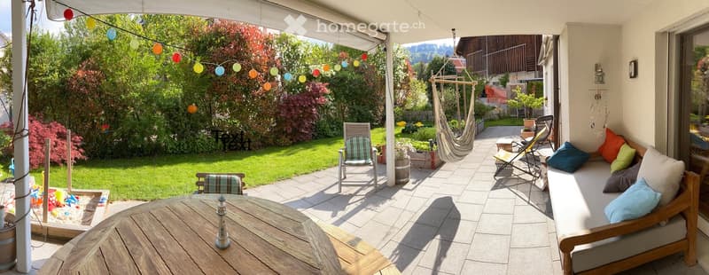 A sunny outdoor patio with a wooden table, lounge chairs, a hammock, and colorful string lights. The area is surrounded by a well-maintained garden with green grass and various plants.