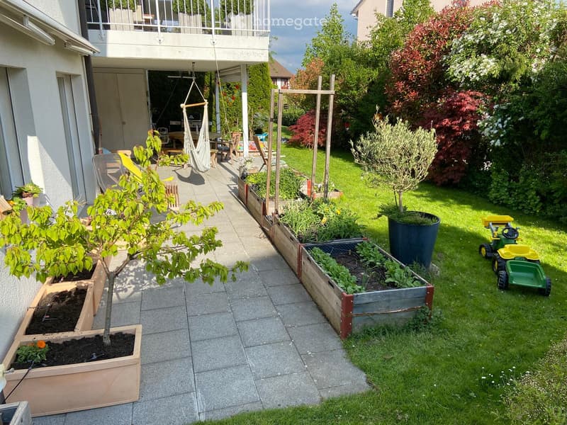 Well-maintained garden with raised wooden planters, a small tree, a hammock, outdoor furniture, and a toy tractor on the grass.