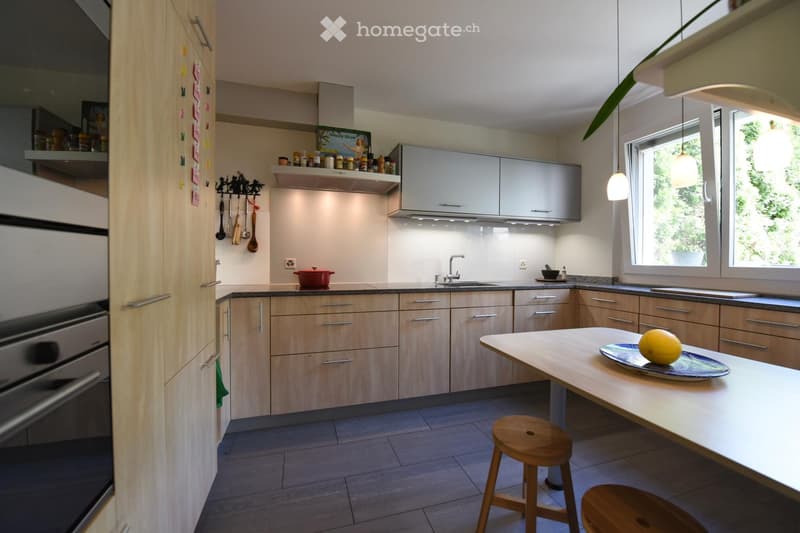 Modern kitchen with light wooden cabinets, stainless steel appliances, and a central island with a stool. Features include a sink, oven, and various kitchen utensils on display.