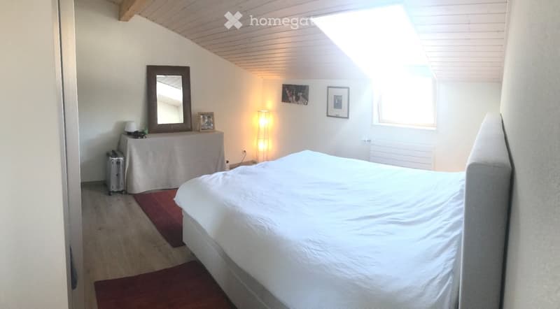 Cozy bedroom with a large bed, a mirror, a small table with a lamp, and framed pictures on the wall. The room features wooden flooring and a slanted ceiling with skylight windows.