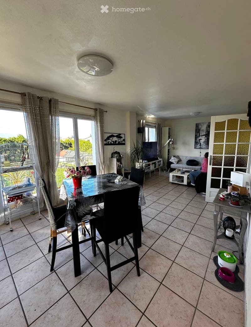 Open-plan living and dining area with tiled flooring, large windows, and a balcony view. Includes a dining table with chairs, a sofa, a television, and decorative items.