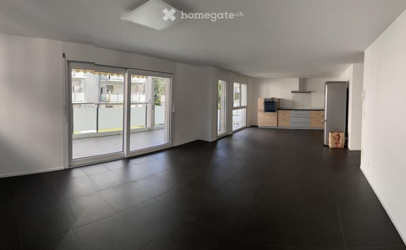 Open-plan unfurnished space with dark tiled flooring, large sliding glass doors leading to a balcony, and a small kitchen area with cabinets and appliances visible in the background.
