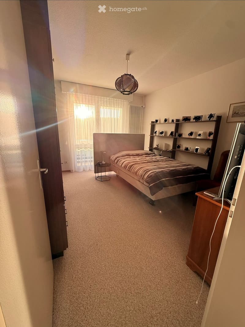 A well-lit bedroom with a bed, nightstand, shelf, chair, and a small table with a lamp. The room features carpeted flooring and large windows with sheer curtains.