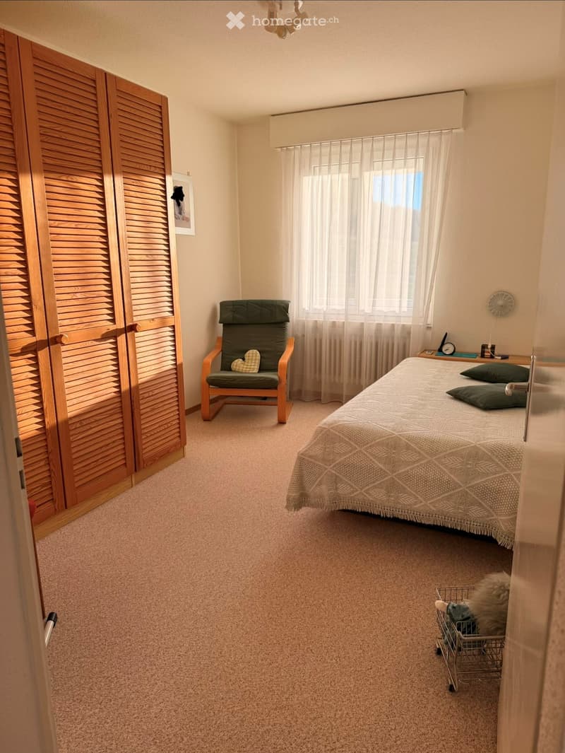 Cozy bedroom with a double bed, green pillows, a wooden wardrobe, a green armchair with a heart-shaped cushion, and a small nightstand with a clock.