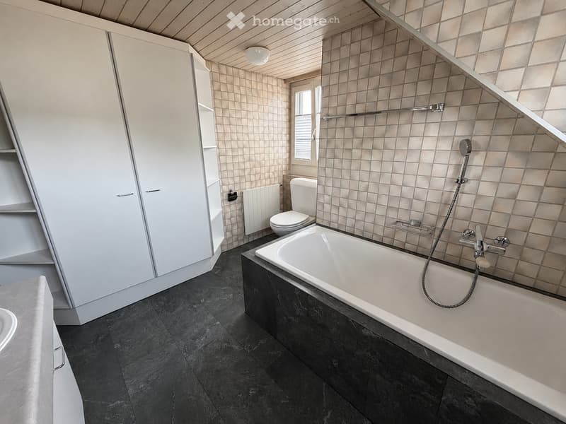 Modern bathroom with a large bathtub, toilet, white cabinet, and tiled walls. Features a sleek, contemporary design with dark flooring and a window providing natural light.