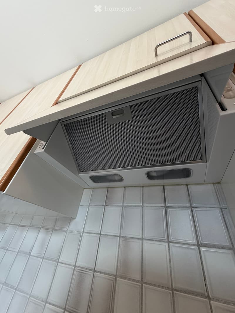 A modern kitchen with light-colored cabinets and a stainless steel range hood, featuring a tiled floor.
