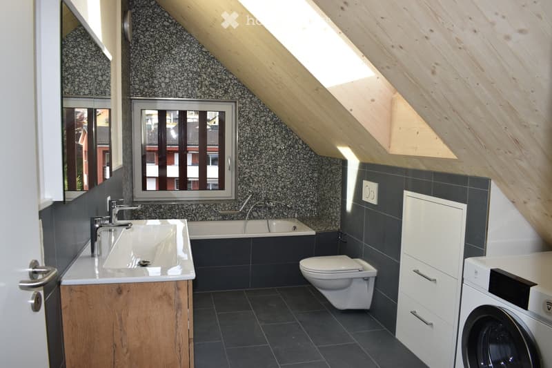 Modern bathroom with a sink, bathtub, toilet, and washing machine. Features include dark tiles, wooden cabinetry, and a large window with a view.