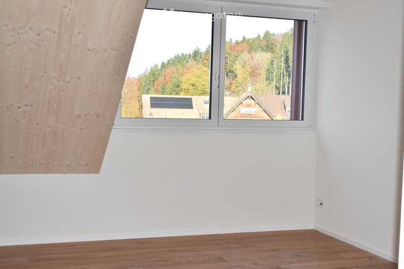 Empty room with white walls, wooden flooring, and large windows offering a view of trees and neighboring houses.