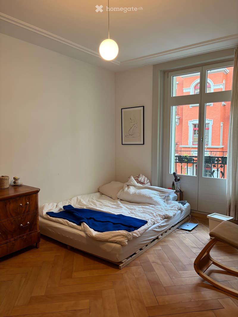 Cozy bedroom with wooden flooring, a single bed with white bedding and a blue blanket, a wooden nightstand, a small table with a cup, a chair, and a picture frame on the wall. A large window with a view of a red building is visible.