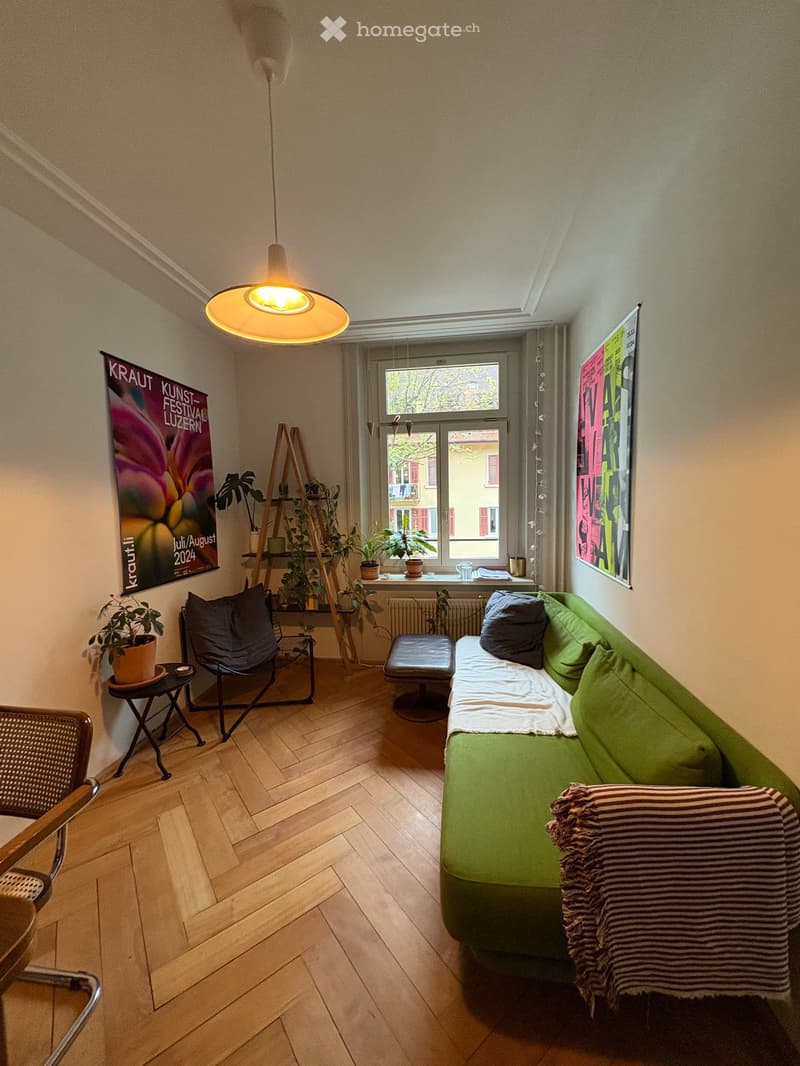 A cozy living room with a green sofa, a black chair, a small table, a stool, potted plants, and posters on the walls. The room features wooden flooring and a hanging lamp.