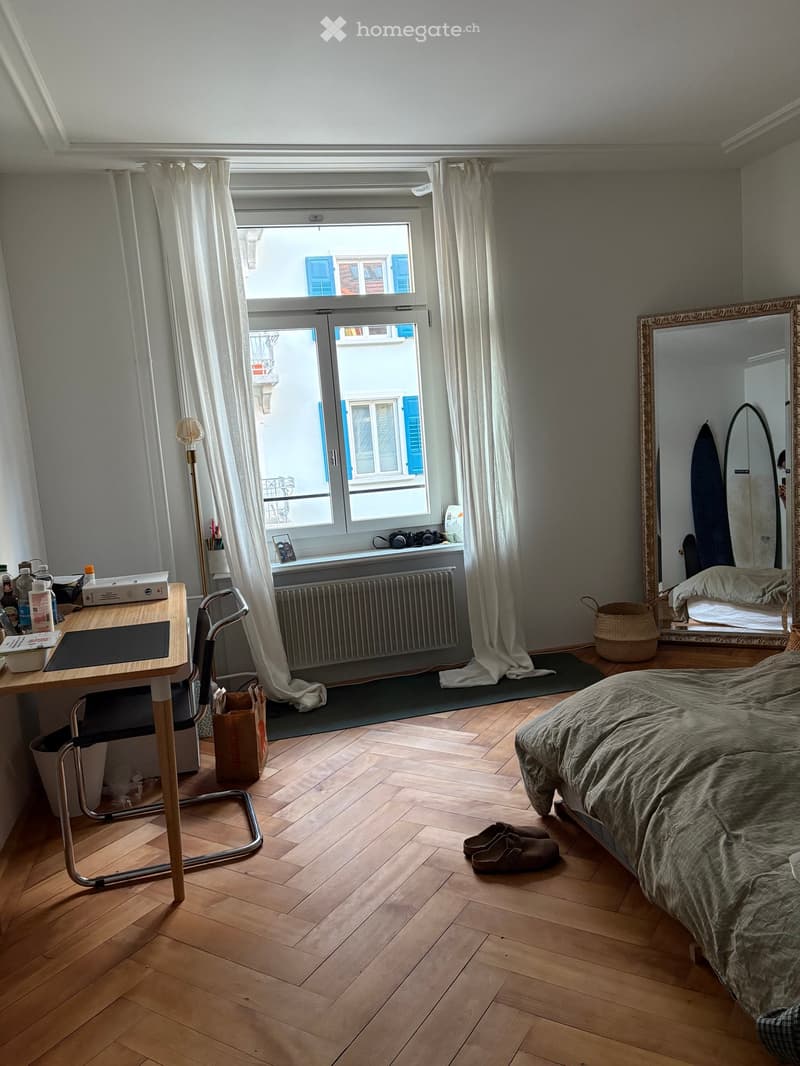 Cozy bedroom with wooden flooring, large window with white curtains, bed with green bedding, desk with chair, mirror, and surfboard in the corner.