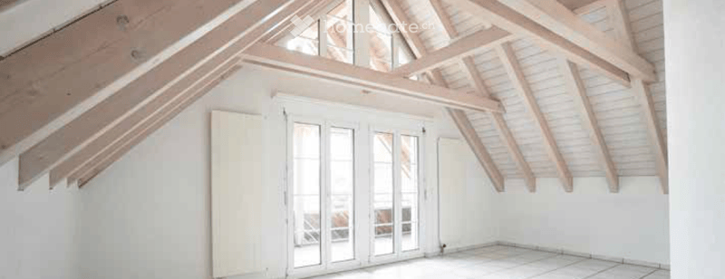 Bright, empty attic space with exposed wooden beams, white walls, and large skylights. The room features a sliding glass door leading to an outdoor area.