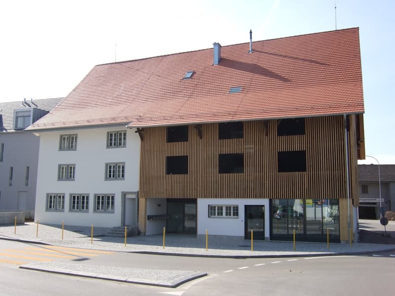 Two-story building with red-tiled roof, white walls, and wooden paneling on one side. Features multiple windows, a glass entrance, and a small staircase leading to the entrance.