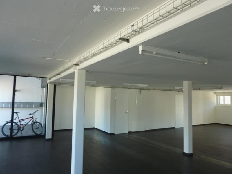 Empty room with dark wooden flooring, white walls, and exposed ceiling beams. A bicycle is visible through a large glass door.