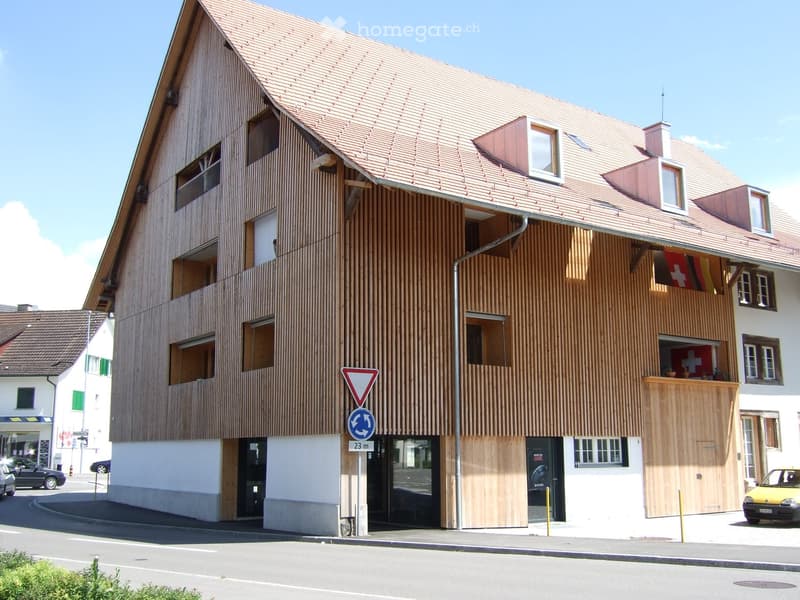 Modern multi-story building with wooden facade, large windows, and a red-tiled roof. Features a balcony with a Swiss flag and a small garden area.