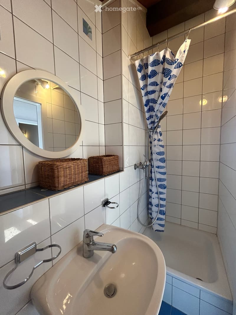 Bathroom with white tiled walls, a sink with a modern faucet, a bathtub with a blue fish-patterned shower curtain, a round mirror, and two wicker baskets on a shelf.