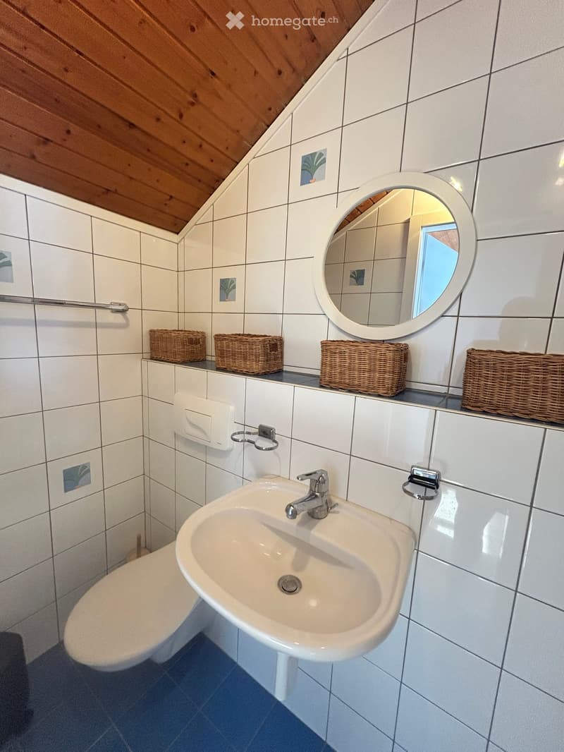 Bathroom with white tiled walls, a sink with a chrome faucet, a toilet, a round mirror, and wicker baskets on the counter.
