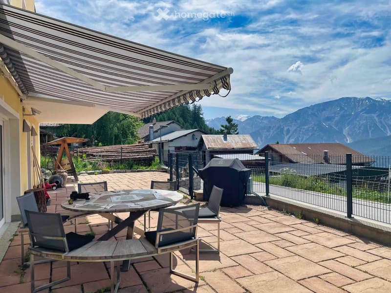 Balcony with a covered awning, a round table with chairs, and a grill, overlooking a scenic mountain view.