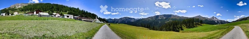 A scenic mountain valley with a winding road, green fields, scattered houses, and distant peaks under a clear blue sky.