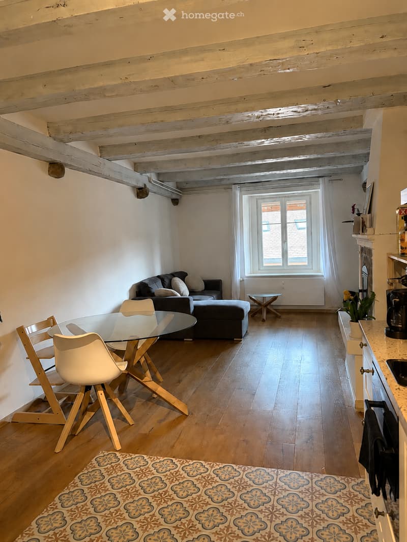 Open-plan living and dining area with exposed wooden beams, hardwood flooring, a grey sofa with ottoman, a glass dining table with four chairs, a small side table, and a kitchen counter with a plant and cup.