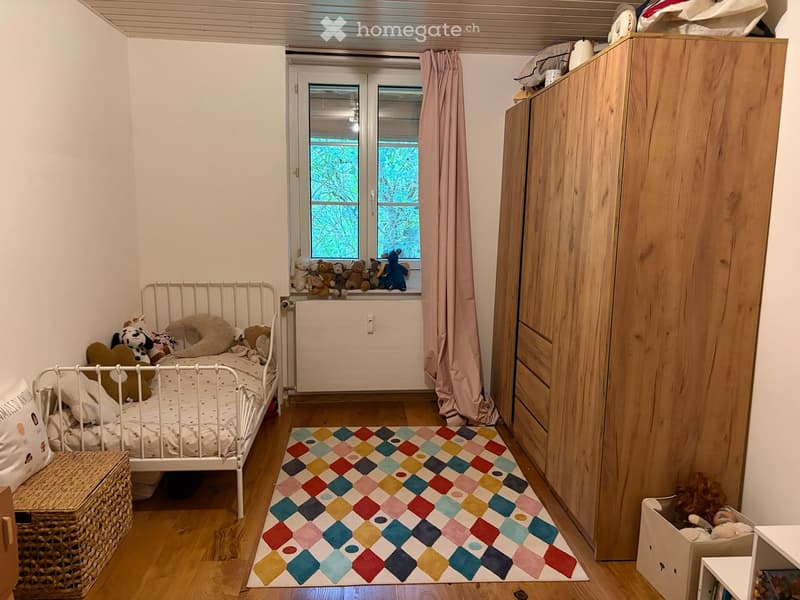 Cozy children's bedroom with a white metal bed, colorful rug, wooden wardrobe, and stuffed toys on the windowsill and bed.