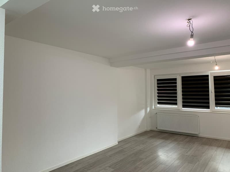 Empty room with white walls, wooden flooring, and large windows with blinds. Features a radiator and ceiling lights.