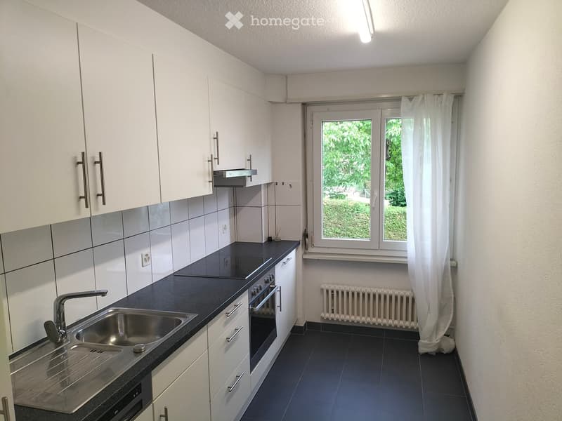 Modern kitchen with white cabinets, black countertop, stainless steel sink, oven, and radiator under window with white curtain.