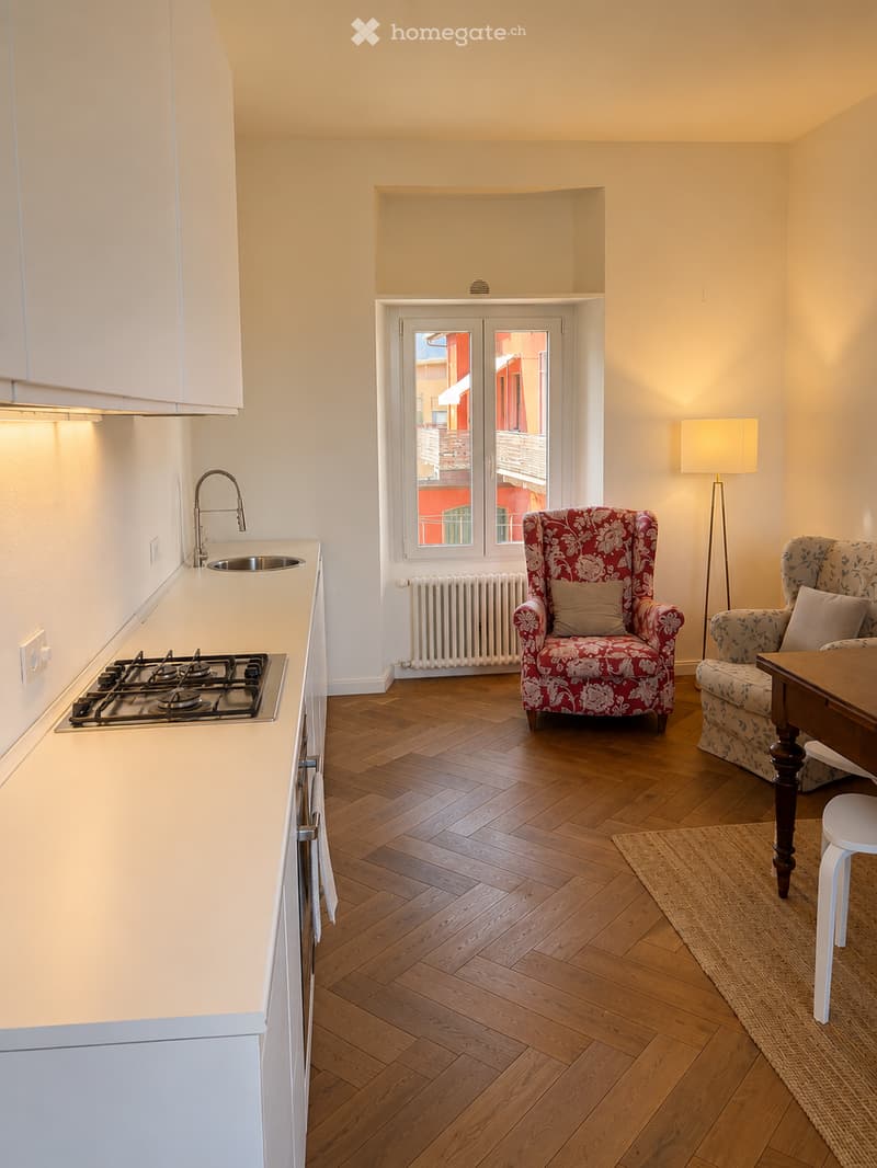 A modern, open-plan living and kitchen area with light-colored cabinets, a gas stove, a sink, and a window with a view. The living area features a patterned sofa, a wooden table, and a floor lamp.