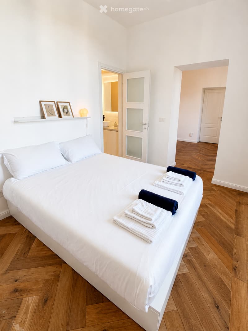 A neatly made double bed with white linens and neatly folded towels, a nightlight, and framed pictures on a shelf in the background.