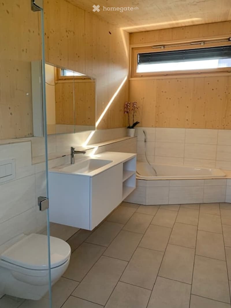 Modern bathroom with wooden walls, white tiles, a large mirror, a sink with a faucet, a bathtub, and a toilet. A small flower vase is placed on the windowsill.