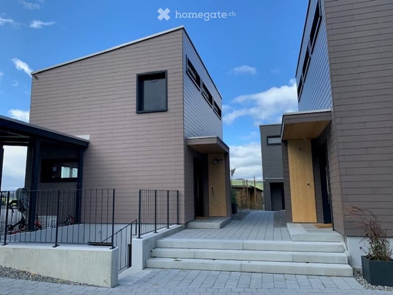 Modern residential building with clean lines, grey siding, and multiple windows. Features a small porch area with steps leading up to the entrance.