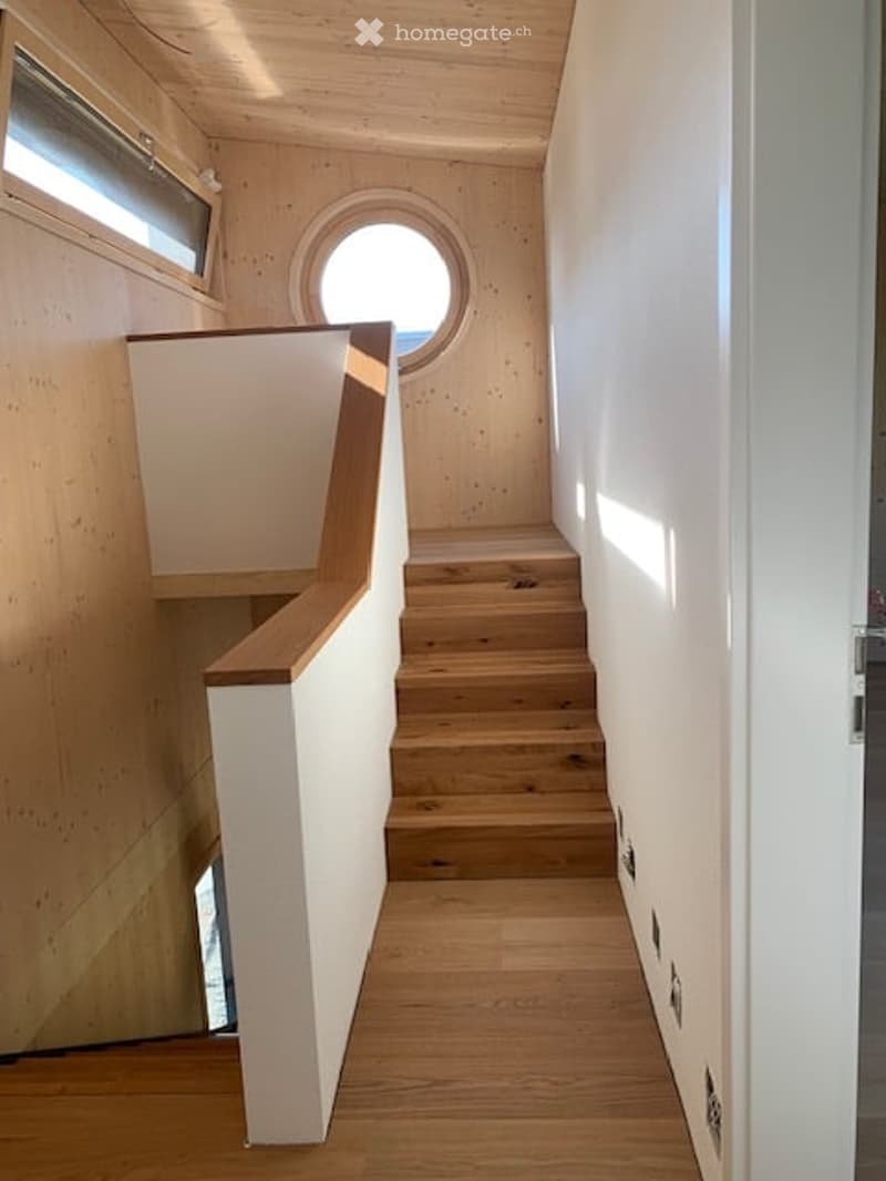 Modern wooden staircase with white walls and circular window, leading to an upper level.