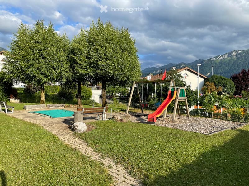 A well-maintained garden area with a swing set, slide, benches, and a grassy lawn, surrounded by trees and a mountainous backdrop.