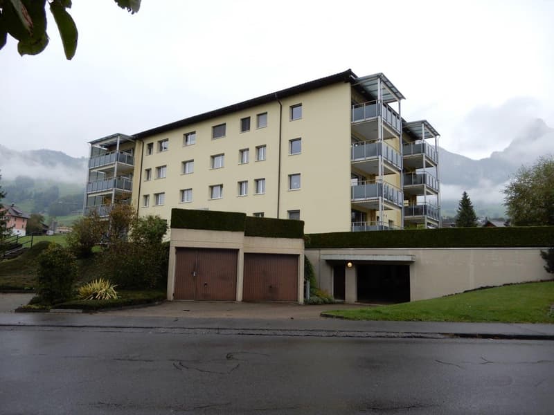Modern multi-story residential building with balconies, garage, and surrounding greenery.