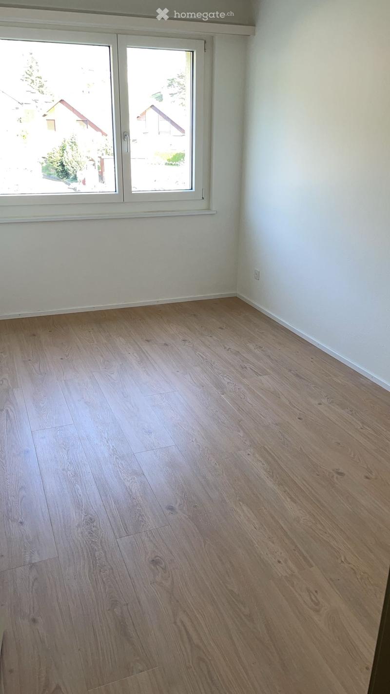 Empty room with light wooden flooring and white walls, featuring a large window with white frames that offers a view of the outside.