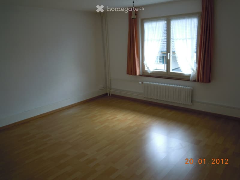 Empty room with wooden flooring, white walls, a window with curtains, and a radiator below the window.