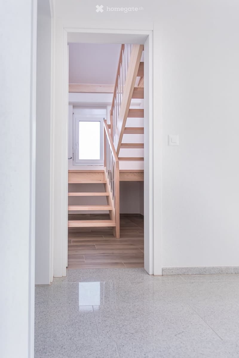 A modern, minimalist hallway with a wooden staircase leading upwards, featuring light-colored wood steps and railings, white walls, and a polished concrete floor.