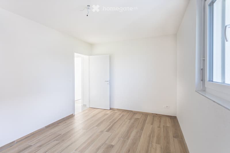 Empty room with light wooden flooring, white walls, and a single door leading to another space. The room has a large window on one side, allowing natural light to enter.