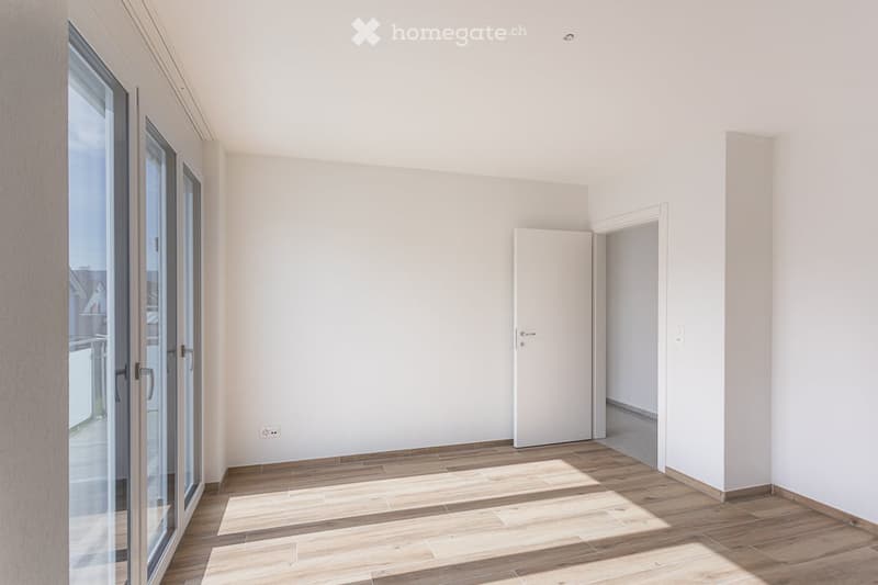 Empty room with light-colored walls, wooden flooring, and large glass doors leading to an outdoor area.
